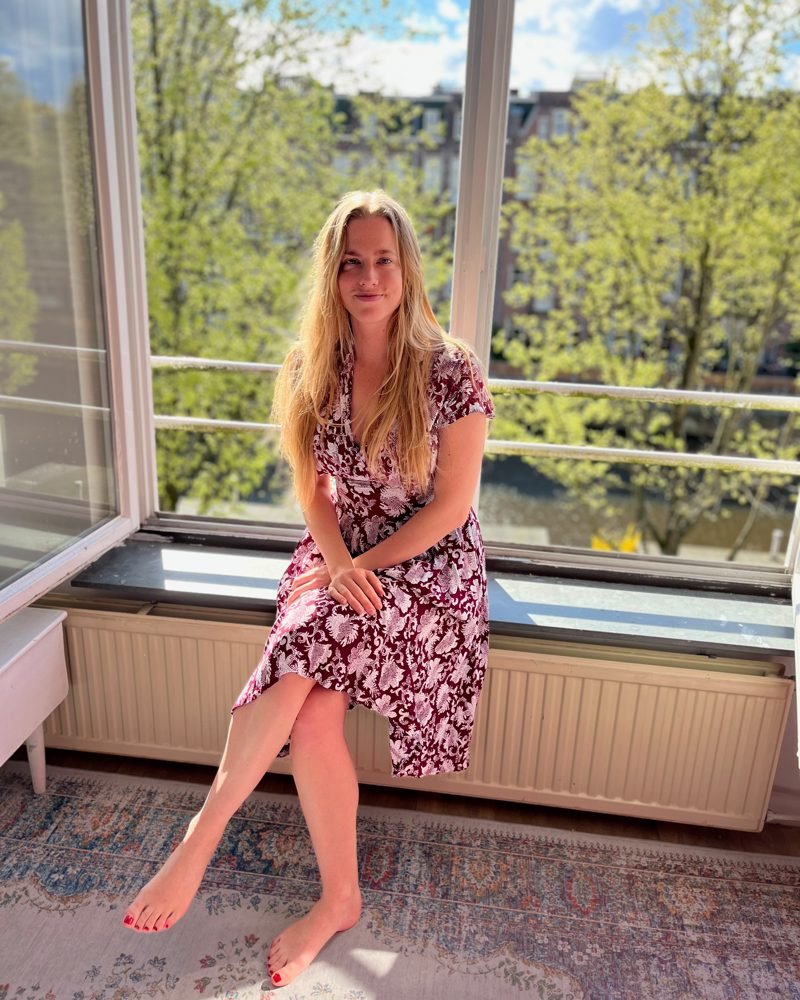 Marte van Os zit in de vensterbank van een open raam, met zon en bomen achter haar